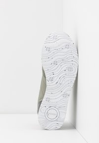 Baskets grises avec une tige texturée, dotée d'une semelle en caoutchouc blanche ondulée avec des taches noires et le logo de la marque. Profil latéral sur fond blanc.