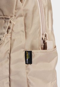 Beige Cordura fabric backpack close-up, featuring a zipper, stitched accents, and a label indicating eco-friendly material.