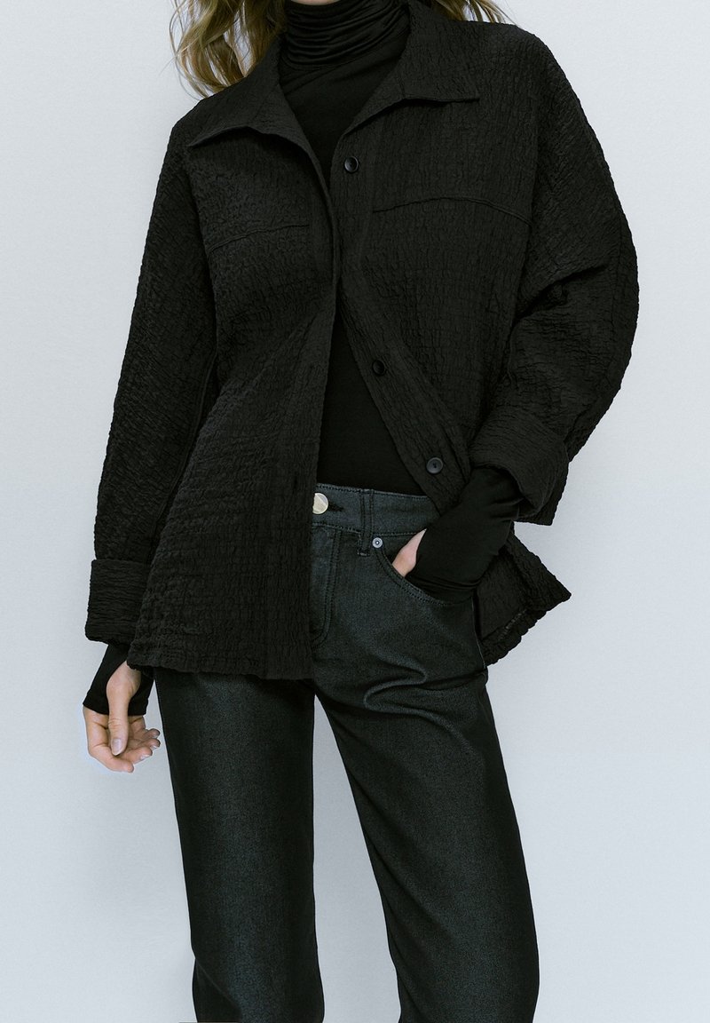Femme portant une veste noire texturée oversize boutonnée sur un col roulé noir avec un pantalon noir, main dans la poche, sur un fond clair uni.