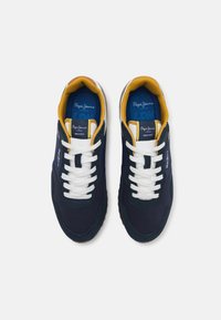 Laivastonsiniset tennarit, joissa on valkoiset nauhat, keltainen sisävuori ja logoyksityiskohta. Näissä kengissä on tekstiili- ja mokkanahkapäällinen, kumipohja sekä klassinen muotoilu.