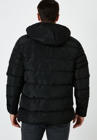 Chaqueta acolchada negra con capucha, con textura acolchada y relleno grueso. La parte trasera muestra una superficie lisa y puños elásticos.