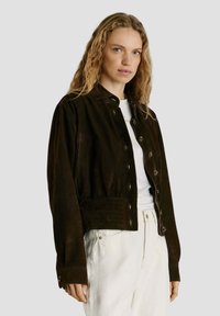 Vaaleat laineikkaat hiukset omaava nainen pukeutuneena tummanruskeaan napitettuun takkiin, jonka alla on valkoinen paita ja luonnonvalkoiset housut. Taustana on yksivärinen vaalea pinta.