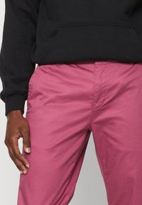 Osoba ubrana w czarną bluzę i jaskrawo różowe spodnie, z jedną ręką swobodnie opartą przy boku na jednolitym tle.