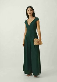 Vestido verde escuro, com mangas em forma de cap sleeve, decote em V, saia rodada e detalhes em pregas; combinado com uma bolsa de mão bege e saltos altos.
