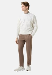 Weißer Pullover mit Zopfmuster, braune, maßgeschneiderte Hose und weiße Turnschuhe. Modell steht seitlich und zeigt strukturierte Details sowie klare Designlinien.