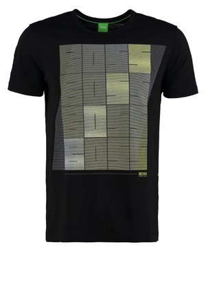 Cardigan noir avec bordure vert citron, fermé par des boutons, porté sur un t-shirt graphique noir, associé à un pantalon noir et des baskets.