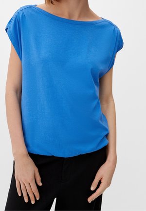 Frau trägt eine hellblaue Kurzarmbluse mit Bootsausschnitt und schwarze Hose vor einfarbigem Hintergrund.