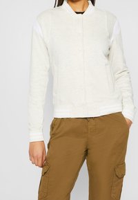 Jasnoszara kurtka bomber z teksturowanej tkaniny, białe akcenty na ramionach, zapięcie na zatrzaski i ściągacze przy mankietach. Połączona z beżowymi spodniami cargo.