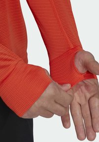 T-shirt de sport à manches longues en orange vif, avec un tissu texturé et des trous pour les pouces dans les poignets pour un ajustement et un confort accrus.