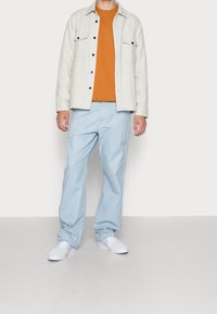 Světlá šedá bunda na knoflíky, oranžové tričko, světle modré kalhoty a bílé boty. Velké kapsy na bundě, uvolněný střih.