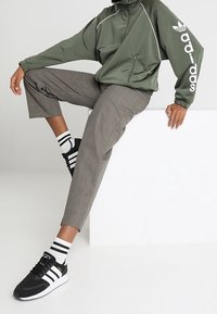 Grön nylonjacka med vita accenter, grårutiga byxor och svarta sneakers med vita ränder. Svartvitrandiga strumpor är synliga.