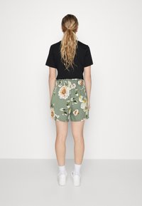 Shorts verdi floreali con una vita elastica, decorati con fiori bianchi e arancioni; abbinati a una maglietta nera e scarpe da ginnastica bianche.