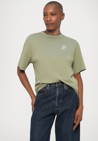 Camiseta de algodón verde claro de mangas cortas, con un pequeño logo blanco en el pecho, combinada con jeans de pierna ancha azul oscuro.