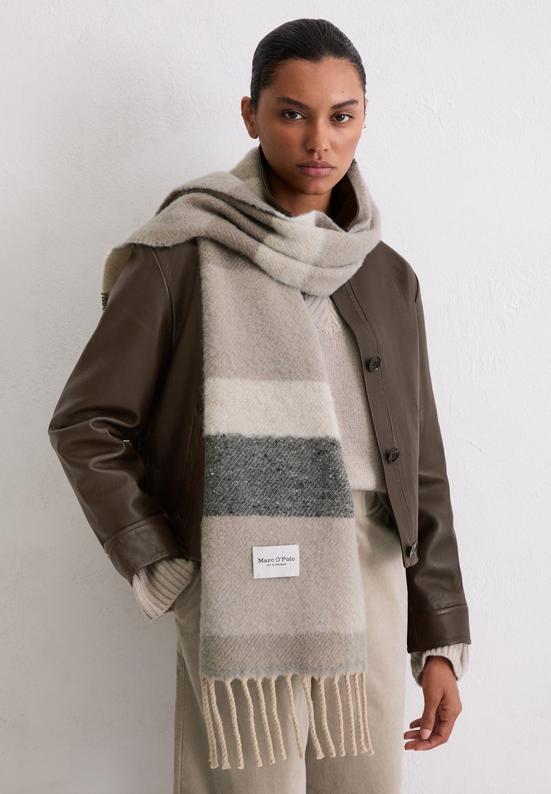 Beżowo-szary, paskowany szalik wykonany z miękkiej wełny, z frędzlami na brzegach i metką z logo, noszony na brązowej skórzanej kurtce.