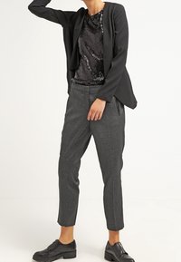 Osoba w czarnym żakiecie, czarnej bluzce z cekinami, szarych dopasowanych spodniach i czarnych skórzanych butach, stojąca z jedną ręką uniesioną do głowy.