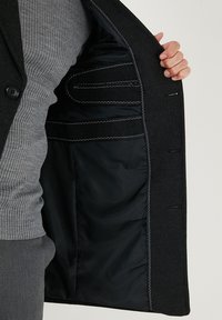 Mustast villasegust bleiserist, millel on sile satiinsisustus, märkimisväärse sisselõikega krae, kaks välimist taskut ja sisemised kohandatud õmblused.