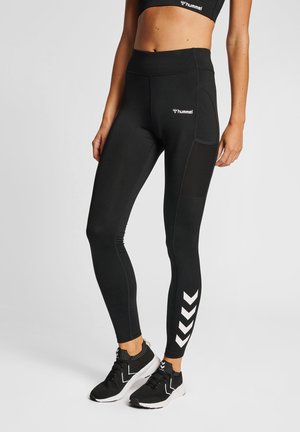 Kvinde iført sorte, højtaljede leggings med sidelomme og hvidt chevron-mønster, parret med sorte sportssko på en grå baggrund.