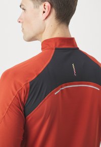 Rot und schwarzes Sportshirt mit hohem Kragen, ausgestattet mit glatter Textur, kontrastierendem Panel-Design und reflektierenden Akzentlinien auf dem Rücken.