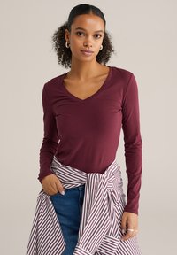Burgundské dlhé rukávy s výstrihom do V v kombinácii s bielou a burgundskou páskovou košeľou viazanou okolo pásu. Hladká textúra, pohodlný strih.