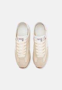 Beżowe zamszowe sneakersy z kremowymi sznurówkami, zakrzywiony design, biały emblemat oraz teksturowana gumowa podeszwa. Widok z góry ukazuje skomplikowane przeszycia.