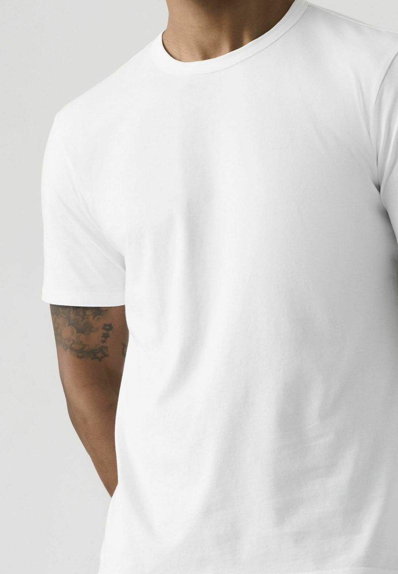 Uomo che indossa una maglietta bianca semplice a maniche corte con scollo rotondo, tatuaggio visibile sull'avambraccio sinistro, su sfondo neutro.