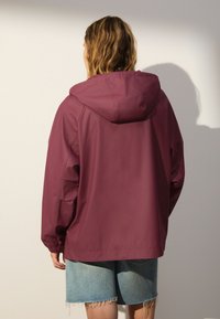 Bordová bunda s kapucňou a elastickými manžetami, s voľným strihom a vodoodolným materiálom, doplnená o roztrhané rifľové krátke nohavice.