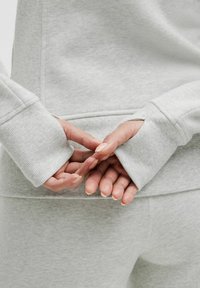 Lys grå sweatshirt med ribbede manchetter, der fremviser en blød stoftekstur. Hænderne er placeret bagpå med fingrene nænsomt flettet.