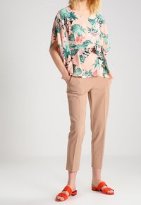 Blusa a fiori rosa con foglie verdi, abbinata a pantaloni beige e sandali scivolati arancioni. Tessuto leggero e fluido, vestibilità rilassata.