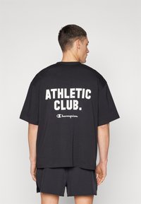 T-shirt en coton noir avec un grand texte blanc "ATHLETIC CLUB." et un plus petit logo "Champion". Coupe décontractée et manches courtes.
