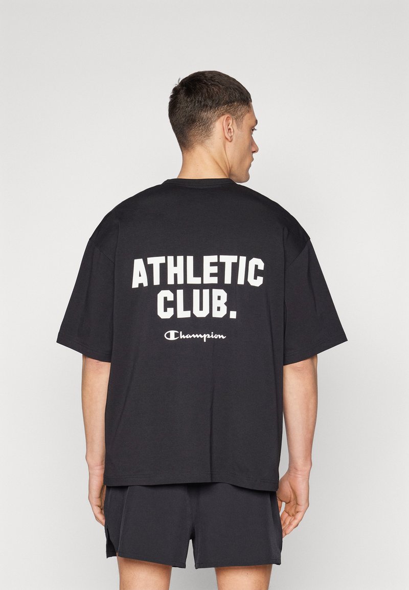 T-shirt en coton noir avec un grand texte blanc "ATHLETIC CLUB." et un plus petit logo "Champion". Coupe décontractée et manches courtes.