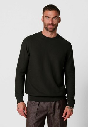 Uomo con capelli castani corti e barba, che indossa un maglione nero semplice a maniche lunghe e pantaloni marroni testurizzati, in piedi davanti a uno sfondo bianco.