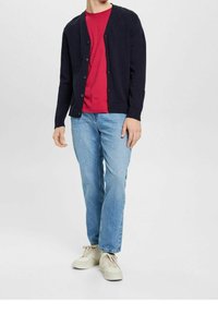 Marinblå stickad cardigan med V-ringning och sex knappar, kombinerad med en röd t-shirt med rund hals och ljusblå raka jeans. Neutrala sneakers.