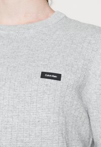Felpa grigia chiara realizzata in tessuto a coste, con un patch del logo Calvin Klein nero sul davanti. Collo a giro classico.