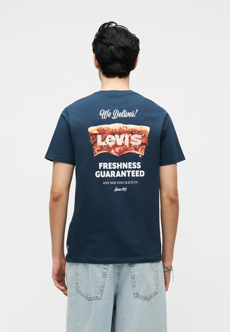 Άνδρας που φοράει μπλε ναυτικό μπλουζάκι Levi's με γραφικό πίτσας και κείμενο "Παρέχουμε παράδοση! Εγγυημένη φρεσκάδα" στο πίσω μέρος, σε συνδυασμό με ανοιχτό μπλε τζιν.