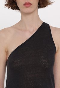 Gros plan d'une personne portant un top noir à une épaule, avec des cheveux courts bruns et un maquillage naturel, sur fond blanc.
