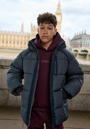 Nastolatek z kręconymi włosami, ubrany w ciemną kurtkę puchową i bordową bluzę z kapturem, stoi nad rzeką, w tle Big Ben i architektura Londynu.