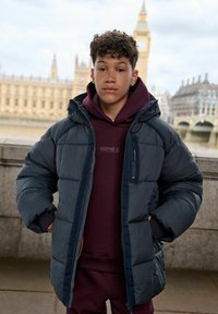 Dunjakke i mørkegrå med hætte, med quiltet design og en frontlynt. Båret over en bordeaux hættetrøje og matchende sweatpants.