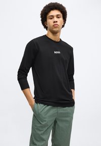 Cămașă neagră cu mâneci lungi, cu logo-ul alb "BOSS" în centru, asortată cu pantaloni verzi în carouri pentru un aspect casual.