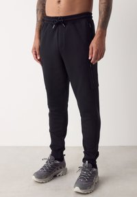 Zwarte atletische joggers met een trekkoord in de taille, zijritszakken en subtiele stikdetails. Gecombineerd met grijze sneakers.