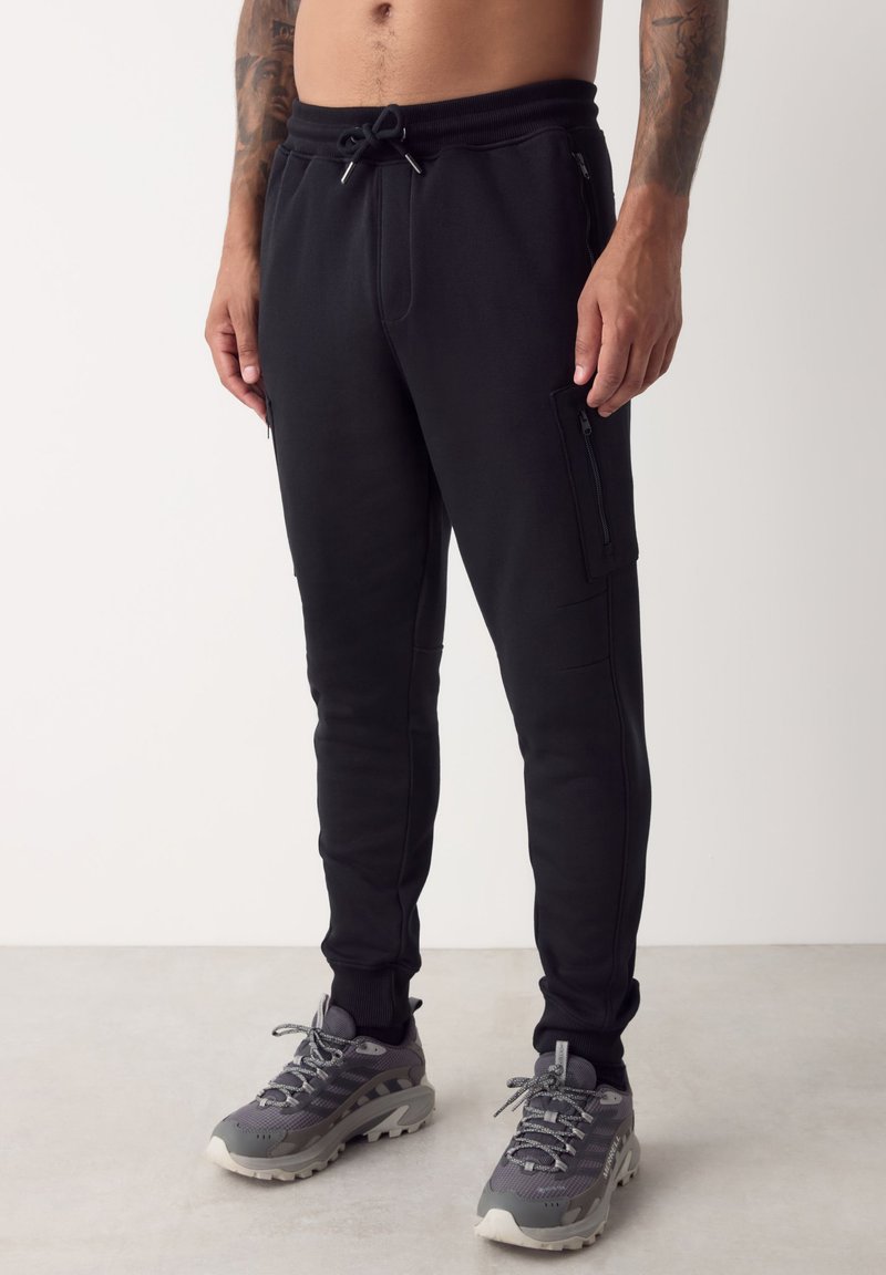 Zwarte atletische joggers met een trekkoord in de taille, zijritszakken en subtiele stikdetails. Gecombineerd met grijze sneakers.