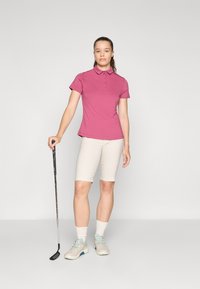 Golf odijelo koje se sastoji od ružičaste polo majice, bež šortsova i svijetlih golf cipela. Majica ima ovratnik i detalje s gumbima.