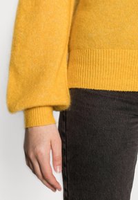 La main gauche de la personne pend à côté d'un pantalon noir, portant un pull jaune à manches longues avec des poignets et une taille côtelés.
