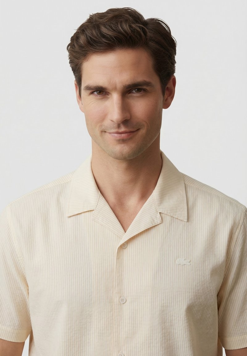 Homme aux cheveux courts et bruns portant une chemise beige clair à manches courtes avec un petit logo crocodile sur la poitrine.