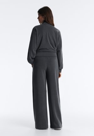 Ženska stoji obrnjena stran, oblečena v temno sivo rebrasto majico z dolgimi rokavi in široke hlače na enobarvnem belem ozadju.