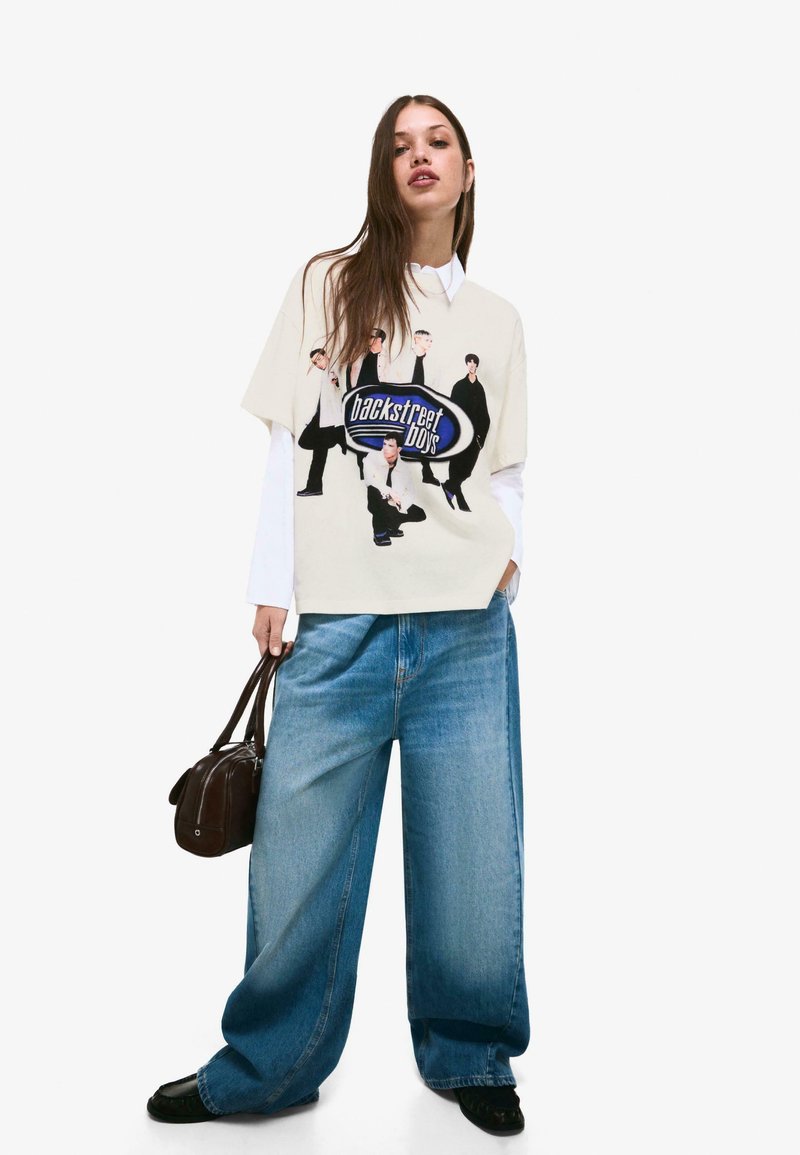 Kremowa, oversize'owa koszulka z grafiką Backstreet Boys, w połączeniu z szerokimi niebieskimi dżinsami. Modelka trzyma małą brązową torebkę.