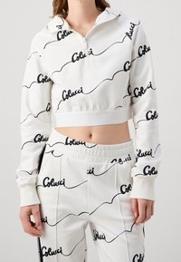 Femme portant un hoodie cropped blanc et un pantalon avec un motif en script noir "Colucci" sur toute la surface, se tenant contre un fond uni.