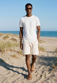 Homme portant des lunettes de soleil, un t-shirt blanc, un short et des sandales marchant sur une plage de sable avec de l’herbe et l’océan en arrière-plan.