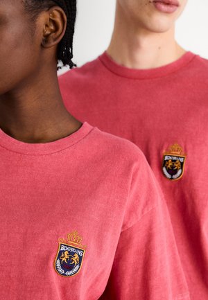 Roze katoenen t-shirts met een ronde hals en korte mouwen, met geborduurde logo's op de borst. De stof heeft een zachte textuur.
