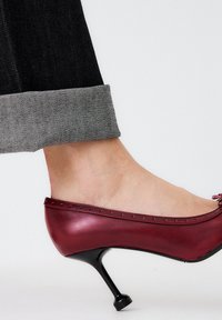 Zapato de tacón alto de cuero rojo con punta redondeada, costuras decorativas y un delgado tacón de aguja negro, combinado con denim oscuro con dobladillos.