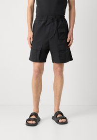 Shorts cargo noirs avec une ceinture élastique, plusieurs poches à rabat et un tissu léger ; associés à des sandales noires.